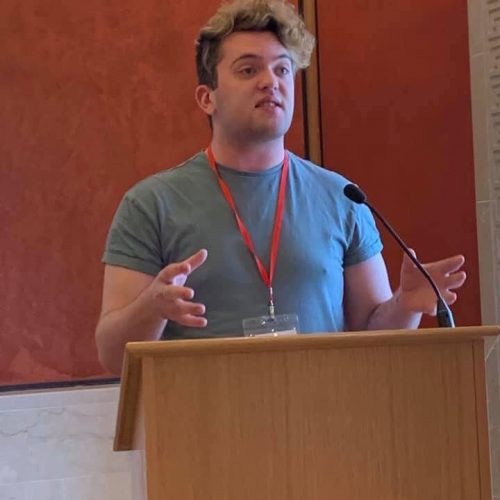 A person stands at a lectern. They are wearing a green t-shirt and speaking at a conference.
