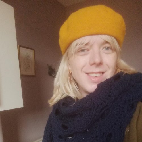 A person wearing a yellow beret and a woolly scarf smiles at the camera.