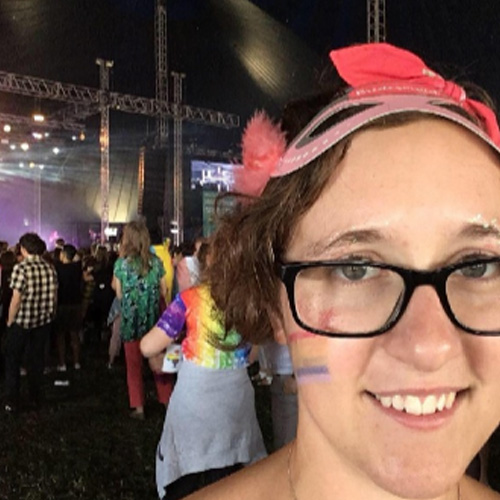 A smiling person with short hair stands close to the camera. They are wearing glasses and have a facepainted rainbow on their cheek.