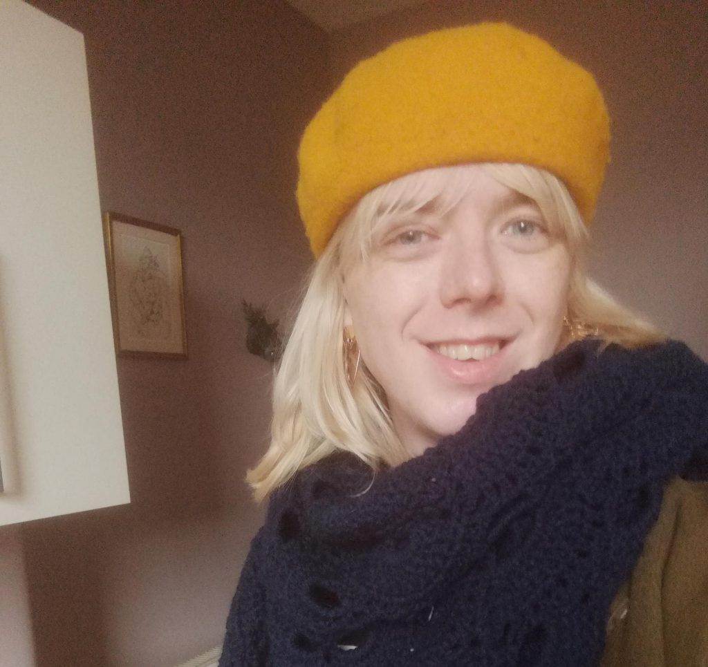 A person wearing a yellow beret and a woolly scarf smiles at the camera.